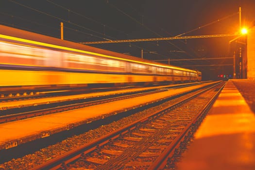 White and blue train passing a small roofless train station at night in the Czech Republic. Blurred motion train.