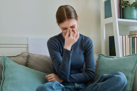 Young suffering teenage female having headache, holding head with hands, sitting on couch at home. Health, problems, mental difficulties, depression, youth concept