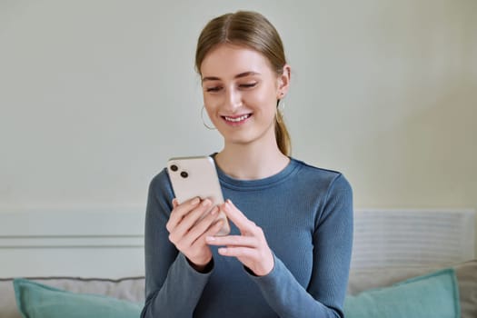 Happy smiling female teenager using smartphone, sitting on couch at home, girl 16,17, 18 years old texting reading messages. Modern digital technologies for communication, leisure, learning, shopping