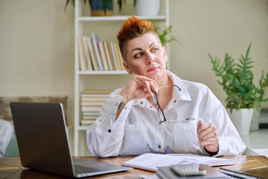 Portrait of mature businesswoman working at home on computer laptop. Serious tired middle-aged female holding glasses in hands sitting at desk in home office. Remote business work management marketing