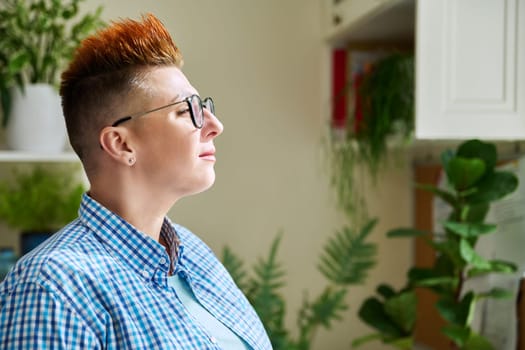 Profile portrait of serious middle-aged woman in glasses with red haircut looking at window in home interior, copy space. Mature people, lifestyle, health, life concept