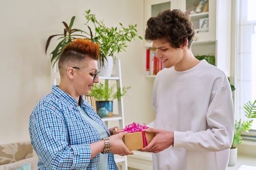 Cheerful teenage son congratulating middle aged mother with surprise gift, standing together at home. Holiday, birthday, congratulations, love, mother's day, happiness, joy, family concept