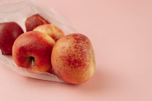 A bag of apples with one of them being slightly bruised. The apples are red and shiny. The bag is white and the background is pink