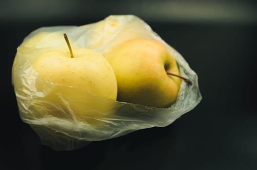 A bag of apples is sitting on a black surface. The apples are yellow and have a stem. The bag is plastic and is placed on a table