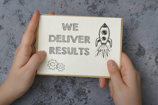 A hand holding a piece of paper with the words We deliver results written on it. The paper also features a rocket on it