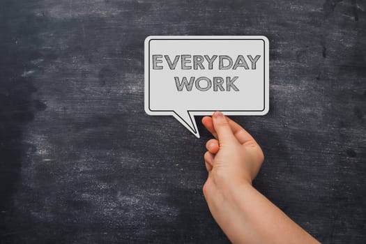 A hand holding a white sign that says everyday work. The sign is on a black background