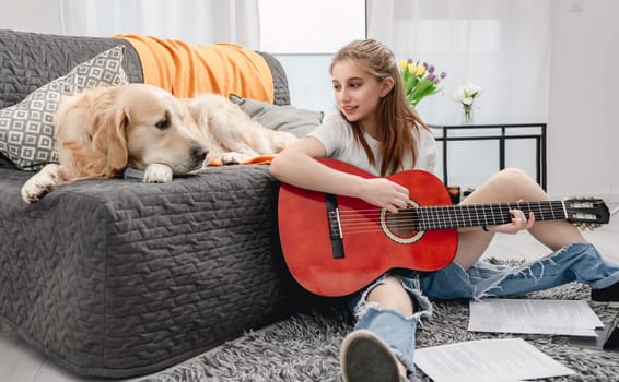 Girl teenager practicing guitar playing with golden retriever dog at home sitting on floor. Pretty guitarist with musician instrument and purebred pet doggy looking at camera