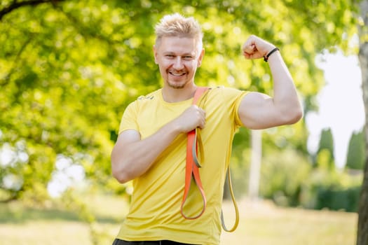 Man with elastic rubber band showing muscles outdoors after arm workout. Guy with sport equipment looking at camera and smiling
