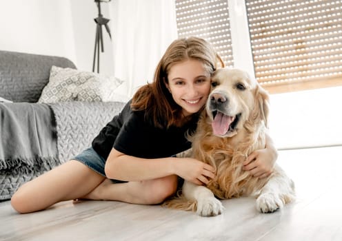 Pretty girl with golden retriever dog sitting on floor at home and hugging pet friend. Beautiful female teenager person with doggy in light room