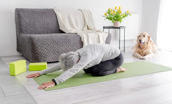 Grey-Haired Woman In baby Pose Practices Yoga At Home On The Floor In A Bright Living Room