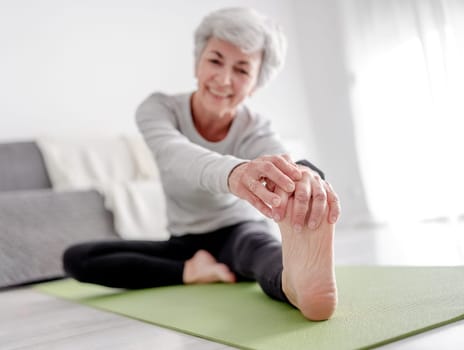 Elderly Athletic Woman Stretches To Her Foot While Sitting On Yoga Mat, Exercising At Home On Floor In Bright Room With Flowers