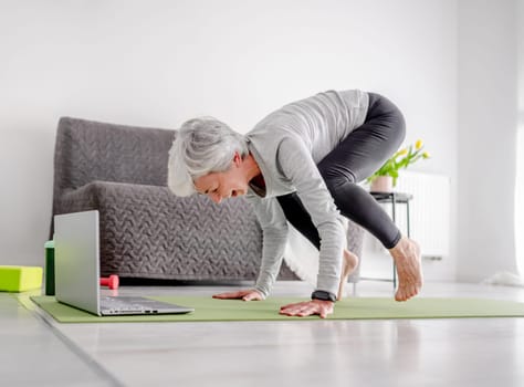 Pleasant 70-Year-Old Woman Performs Complex Exercises With Trainer Through Laptop, Doing Yoga Online At Home