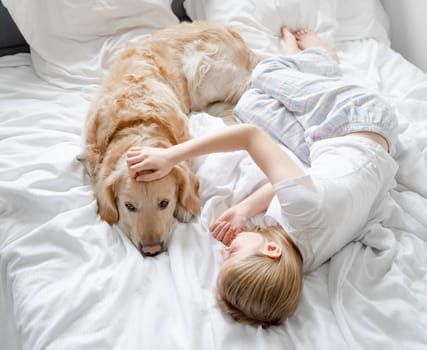 Small Girl Lies On Bed Playing With Golden Retriever Dog On White Bedding