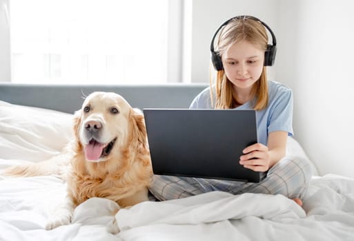 Girl Sits In Bed With Laptop, Headphones And Golden Retriever Dog In Bright Room In The Morning