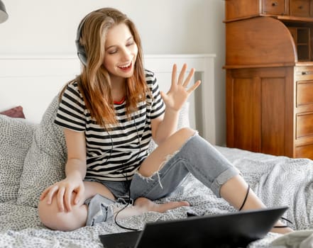 Pretty girl with headphones and laptop in bed during online learning webinar. Beautiful teenager talking by video call with pc computer at bedroom