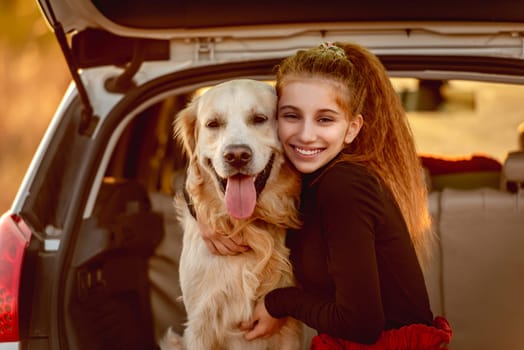 Smiling teenage girl hugging golden retriever dog in car trunk under sunlight