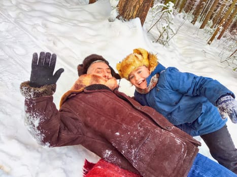 Father and son hugging, having fun, walking in snow nature. Photo shoot in stylized clothes of the USSR. Hat with earflaps. Happy dad and teenager child playing in winter park, forest in holidays