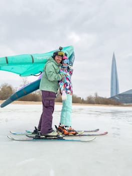 Russia, St.Petersburg, 14 March 2024: A happy couple in bright colored ski gear are ready to ski on ice with an air wing at cloudy weather, the Lakhta Center skyscraper, helmet and glasses. High quality photo