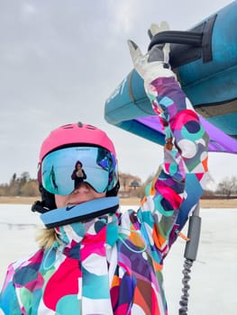 Russia, St.Petersburg, 14 March 2024: A girl in bright colored ski gear is ready to ski on ice with an air wing at cloudy weather, helmet and glasses on her head. High quality photo