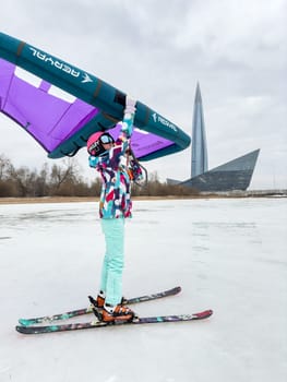 Russia, St.Petersburg, 14 March 2024: A girl in bright colored ski gear is ready to ski on ice with an air wing at cloudy weather, the Lakhta Center skyscraper in the background, helmet and glasses. High quality photo