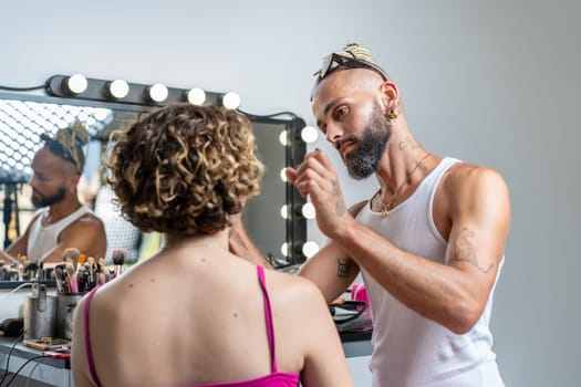 Focused bearded gay professional make-up artist applying makeup on female model with brush sitting in front of mirror in studio