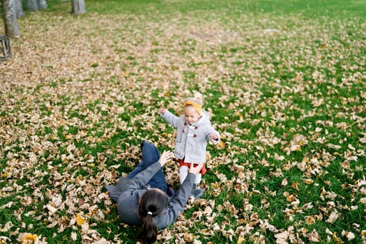 Mom lying on dry foliage stretches out her hands to a little girl standing on a green lawn. High quality photo