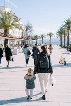 Mother and a little girl are walking holding hands along the embankment with green palm trees. Back view. High quality photo