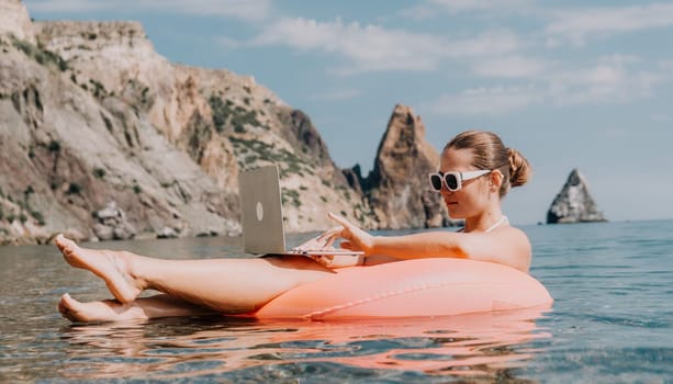 Woman freelancer works on laptop swimming in sea on pink inflatable ring. Pretty lady typing on computer while floating in the sea on inflatable donut at sunset. Freelance, remote work on vacation