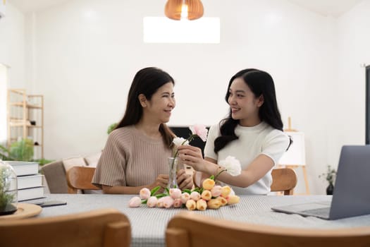 Senior mother and adult daughter happy on the table while arrange flowers in a vase together. Technology and lifestyle concept. Happy time together.