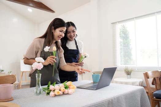 Senior mother and adult daughter happy on the table while arrange flowers in a vase together using laptop computer. lifestyle concept. Happy time together.