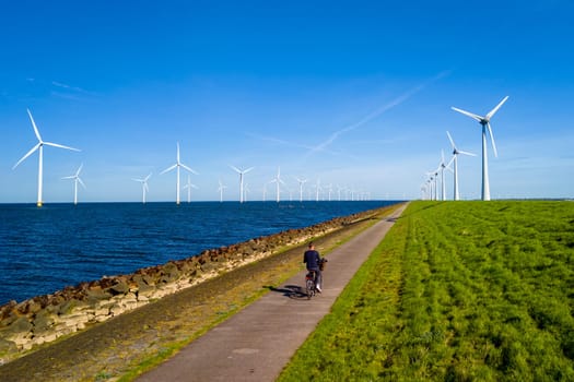 A man riding a bike down a path next to a vast wind farm in the Netherlands Flevoland during Spring, with windmill turbines spinning in the background.