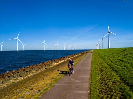 A man cycling along a path beside a serene body of water, with windmill turbines in the distance under the bright Spring sky., men on an electric bike in the countryside in Flevoland Netherlands