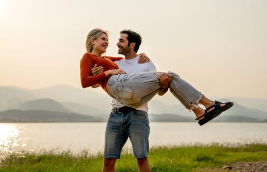 Caucasian man stand and carry his lover woman with love and stay near lake in park with sunset light and they look happiness together.