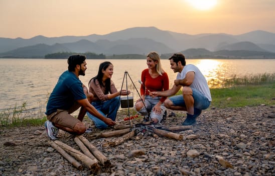 Group of men and woman friends with multi-ethnic enjoy to party and stay surrounded by fire and they look happy to join together in public park near lake and sunset.