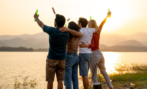 Back of group of friends with multi-ethnic hug together also hold bottle of drinking stand in front of sunset light near lak of public park with happiness.