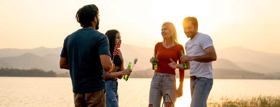 Group of men and women friends with multi-ethnic enjoy with talking and hold bottle of drinking in public park near the lake with sunset light.