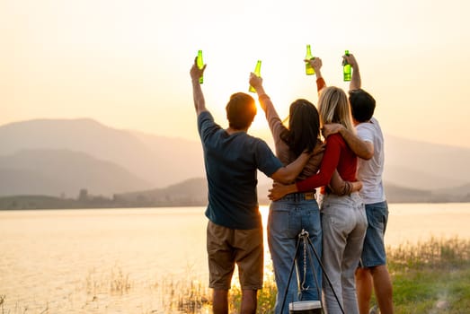 Wide shot of Back of group of friends with multi-ethnic hug together also hold bottle of drinking stand in front of sunset light near lake of public park.