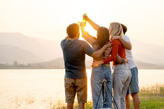 Back of group of friends with multi-ethnic hug together also hold bottle of drinking and cheers action with stand in front of sunset light near lak of public park with happiness.