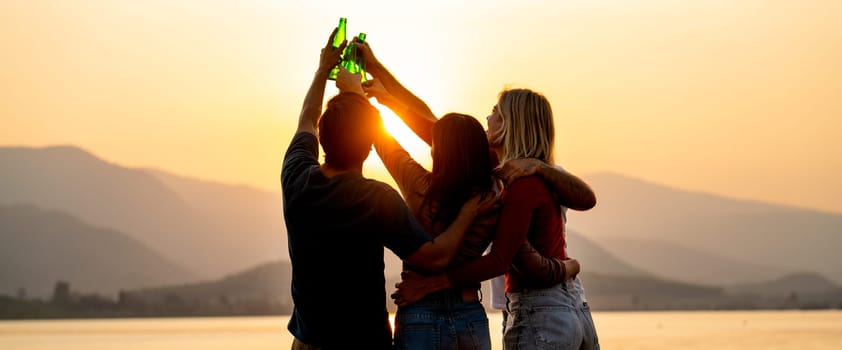 Silhouette of group of friends with multi-ethnic hug together also hold bottle of drinking and cheers action with stand in front of sunset light near lak of public park.