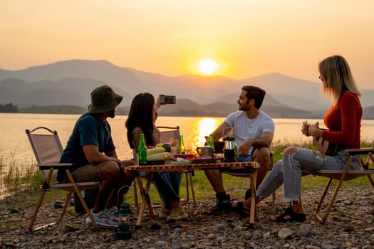 Beautiful Asian woman use mobile phone to selfie with her friends during camping and enjoy together near lake with sunset and they look happy for party outdoor.