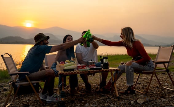 Group of multi-ethnic friends men and women sit on chair with party and camping together also cheers with bottle near lake in public park.