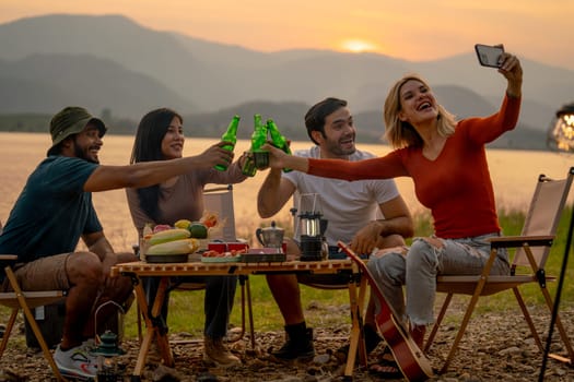 Beautiful Caucasian woman use mobile phone to selfie with her friend during camping and enjoy together near lake with sunset and they look happy for party outdoor.