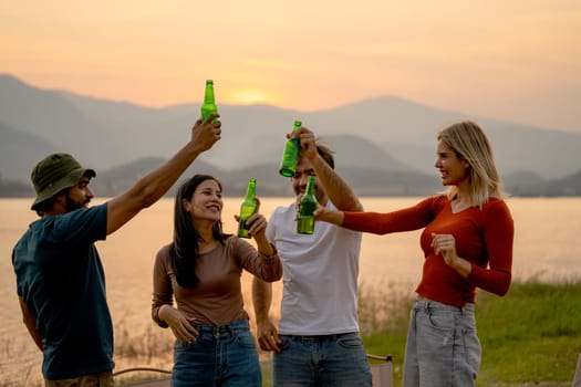 Group of multi-ethnic friends men and women enjoy with small party or camping with hold bottle of drinking near lake in public park and sunset light.