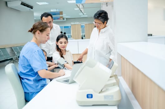 Caucasian nurse measure blood pressure of Asian woman and support by her family with husband, little girl and grandmother in area of hallway of hospital.