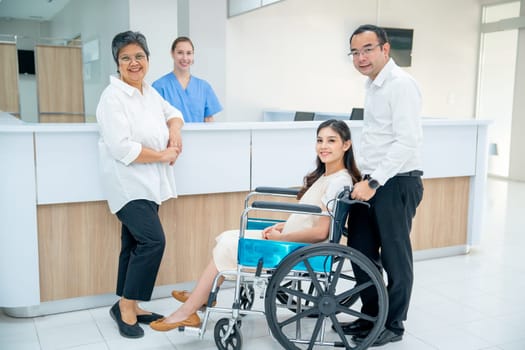 Asian family with senior woman, pregnancy woman sit on wheelchair and man take care the woman, they look at camera with nurse is smiling in the background in area of registration of hospital.