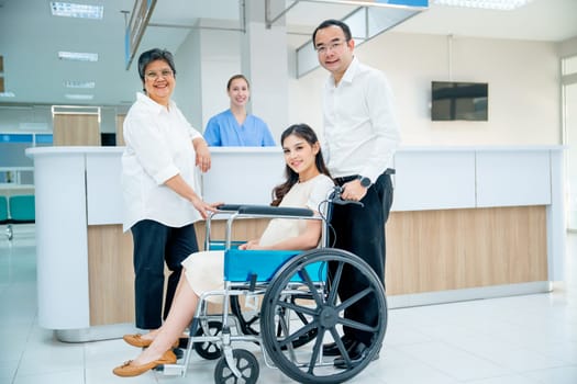 Wide shot of Asian family with senior woman, pregnancy woman sit on wheelchair and man take care the woman, they look at camera with nurse is smiling in the background in area of registration.