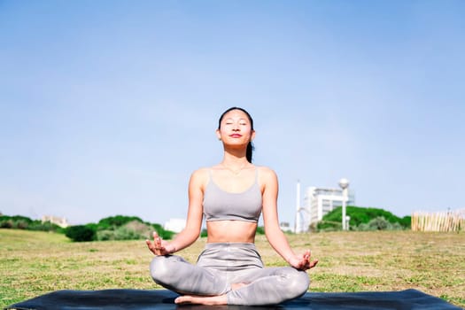 young asian woman doing meditation at park sitting on a yoga mat, concept of mental relaxation and healthy lifestyle, copy space for text