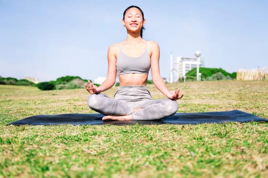 smiling young asian woman doing meditation at park sitting on a yoga mat, concept of mental relaxation and healthy lifestyle, copy space for text