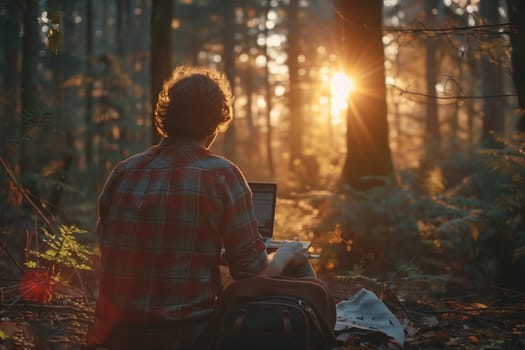 A man sits among the trees in a woodland area, using a laptop computer surrounded by natures beauty. The heat and darkness of the forest create a peaceful atmosphere for his work