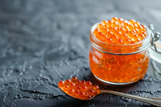 Close up of premium red fish caviar in a glass jar against a dark stone textured background, with a spoonful of caviar delicately placed aside.